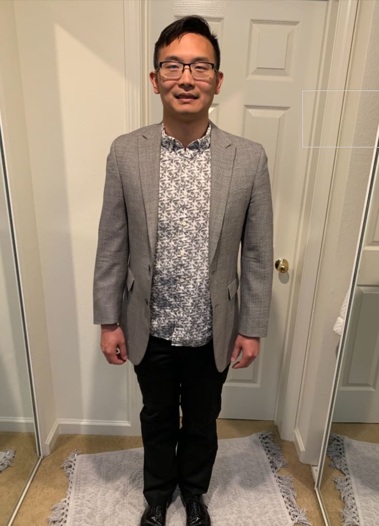 A person in a gray blazer and patterned shirt stands in front of a white door, with mirrors on both sides.
